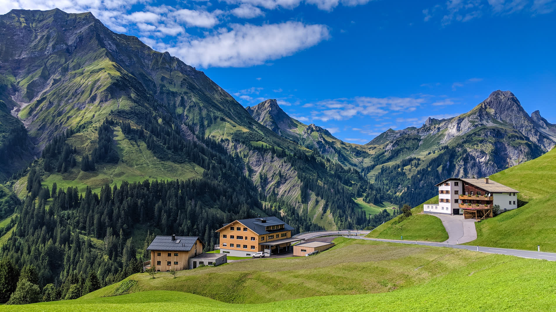 Bergtal mit Häusern und einer Straße, dahinter schroffe Gipfel unter blauem Himmel
