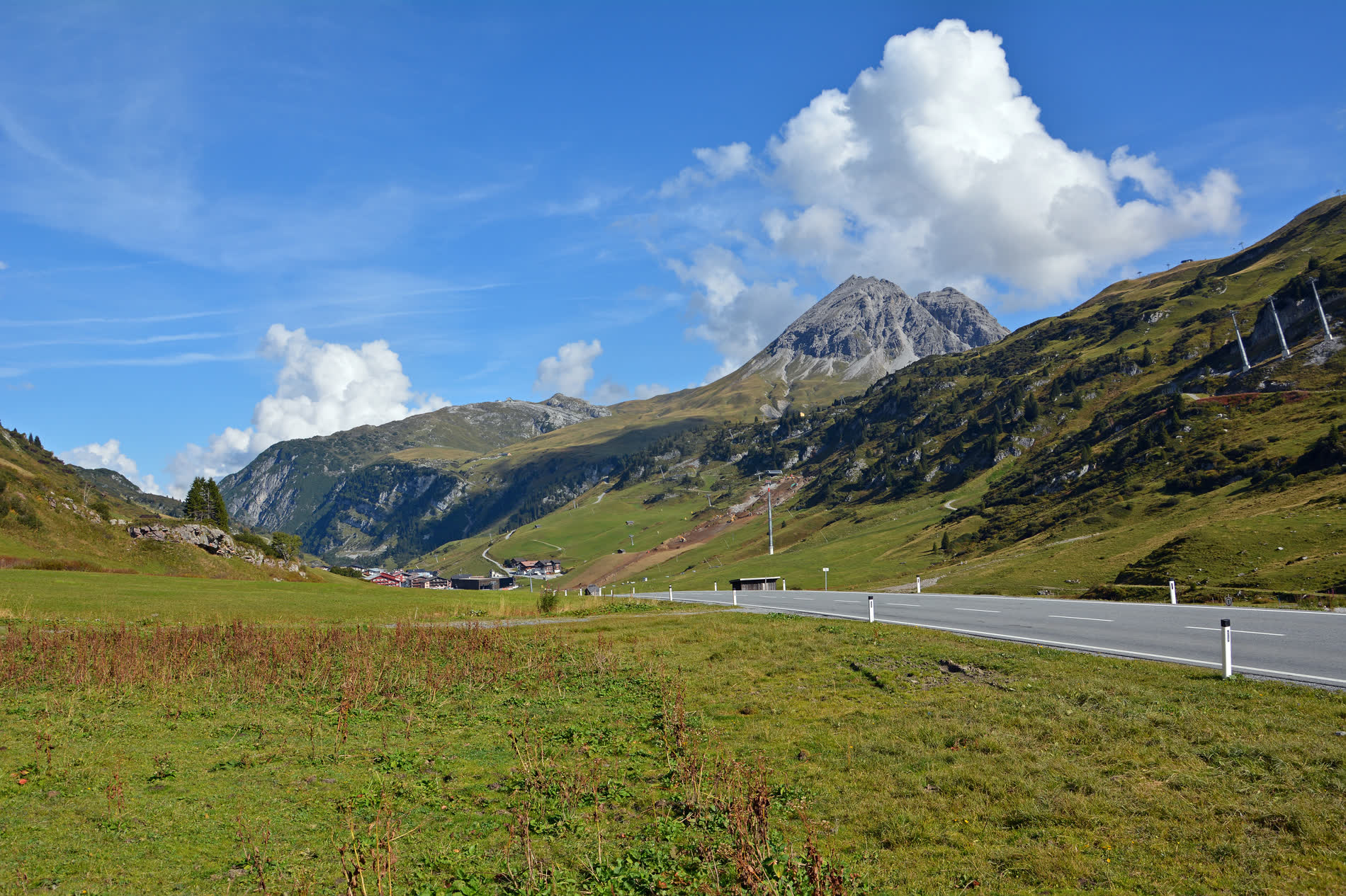 Passstraße durch grüne Almhänge, dahinter ein spitzer Felsgipfel unter Wolken