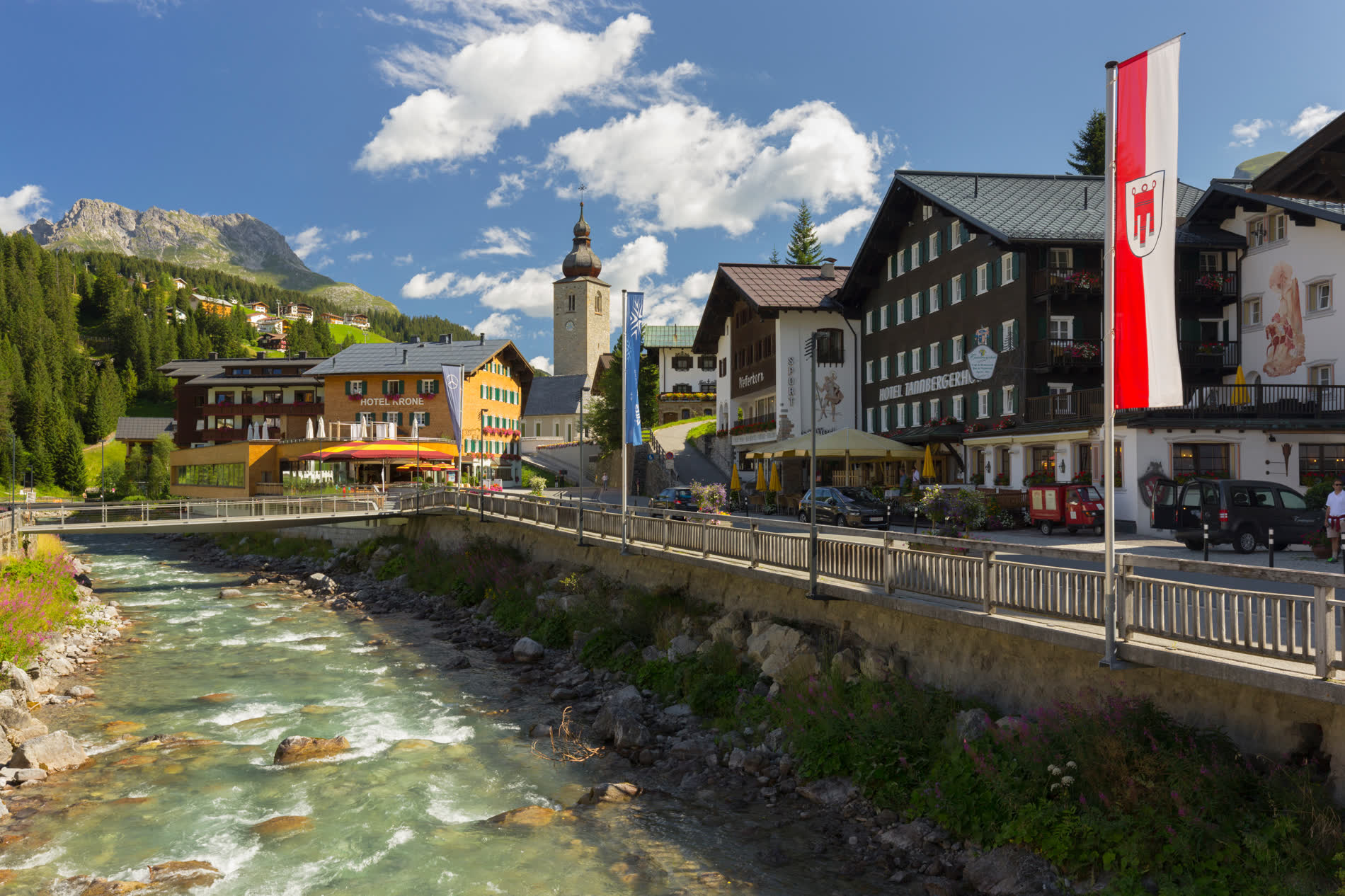 Gebirgsbach unter einer Uferstraße, dahinter Hotels, eine Kirche und eine rot-weiße Fahne